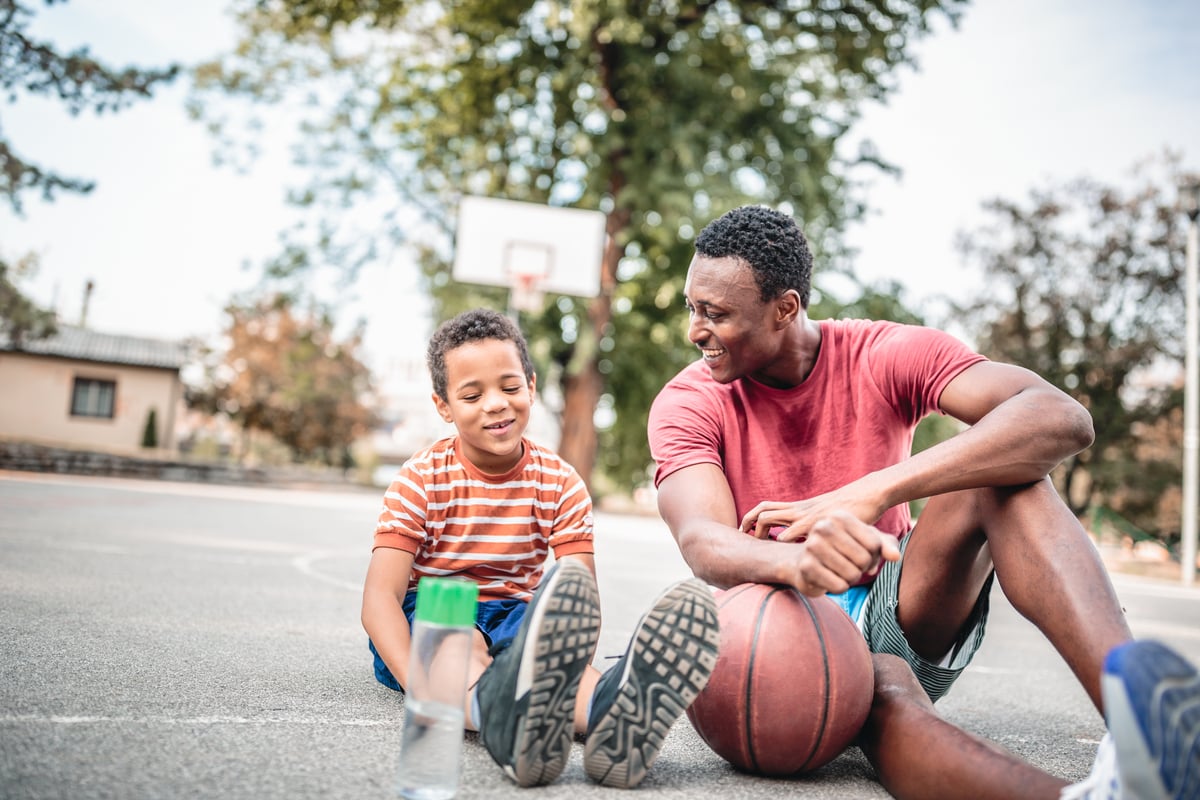 An adult and a child smiling while sitting on a basketball court with a basketball and a water bottle.